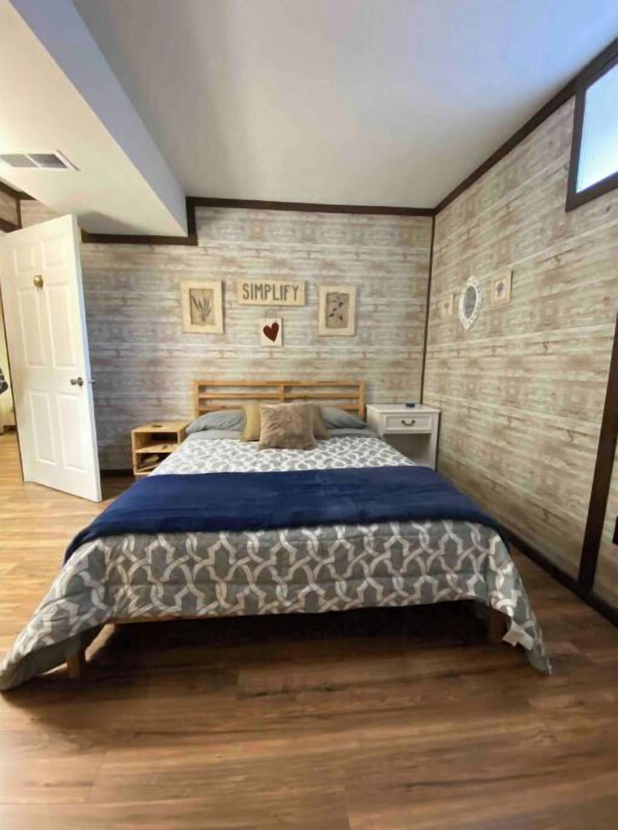 A bedroom with a wooden floor, a bed with a patterned bedspread, a nightstand, and a wall with framed pictures and the word "SIMPLIFY".