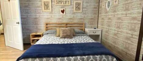 A bedroom with a wooden floor, a bed with a patterned bedspread, a nightstand, and a wall with framed pictures and the word "SIMPLIFY".