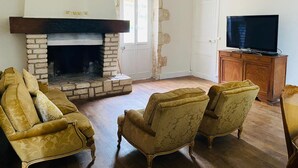 A living room with a stone fireplace, two armchairs, a television on a wooden cabinet, and a door with a stone wall. - Cosy Cottage with pool in the countryside, France (Persac)