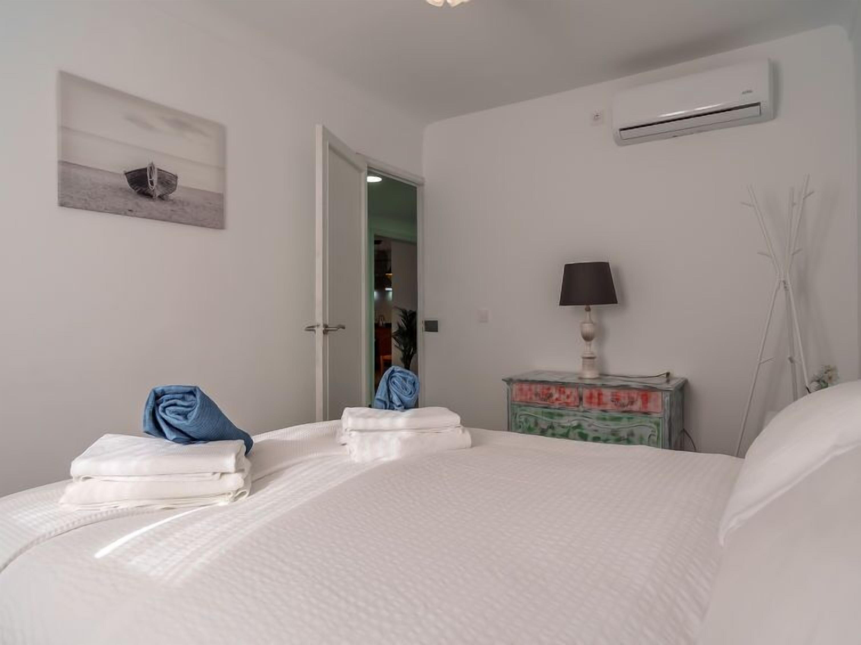 Habitació d'hotel amb un llit, una tauleta de nit amb una làmpada, aire condicionat fixat a la paret i una porta que dona a una altra habitació.