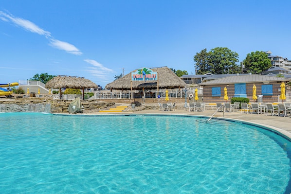A clear blue pool with lounge chairs and umbrellas, a thatched-roof building labeled "TIKI HUT", and a stone wall in the background.