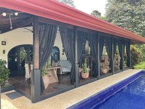 A covered outdoor area with a pool, featuring a covered seating area with curtains, a wooden bench, and potted plants.