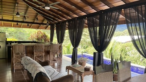 A spacious living area with a wooden ceiling, a sofa, a dining table, and a view of a swimming pool and lush greenery. - VIlla costa pura vida (agalla) (Ojochal)