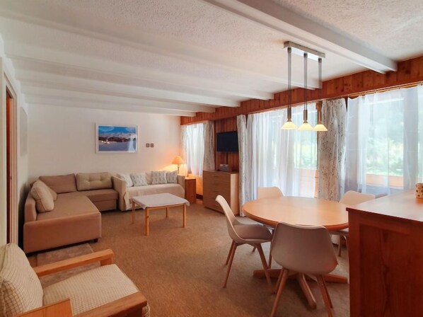 A cozy living room with a sofa, a dining table with chairs, a TV, and a window with curtains.