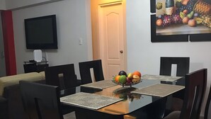 Comedor con una mesa dispuesta para cuatro personas, rodeada de sillas. Un televisor está montado en la pared, y se ve una puerta al fondo.