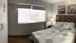 Un dormitorio con cama, mesita de noche, lámpara, ventana con persianas y dos adornos de pared.
