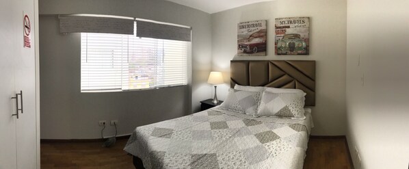 A bedroom with a bed, a nightstand, a lamp, a window with blinds, and two wall decorations. - Charming & Luxury Condo (Miraflores)