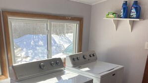 A laundry room with two white washers and dryers, a window with a view of a snowy landscape, and a shelf with cleaning products. - Gateway to the Keweenaw  (Allouez)