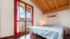 Une chambre avec un plafond en bois, un lit recouvert d’une couvre-lit à motifs floraux, une fenêtre avec des rideaux rouges et un luminaire fixé au mur.