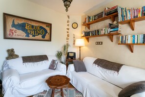 Un salon avec deux canapés blancs, une table basse en bois, une bibliothèque, une horloge et un tableau encadré représentant un rhinocéros bleu.