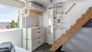Et soveværelse med et vindue, en trætrappe, en hvid kommode og en hylde med toiletartikler.