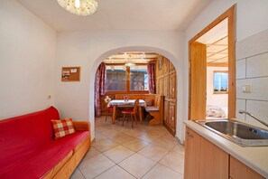 A cozy room with a red sofa, a wooden dining table with chairs, a kitchen area with a sink, and a view through an archway.