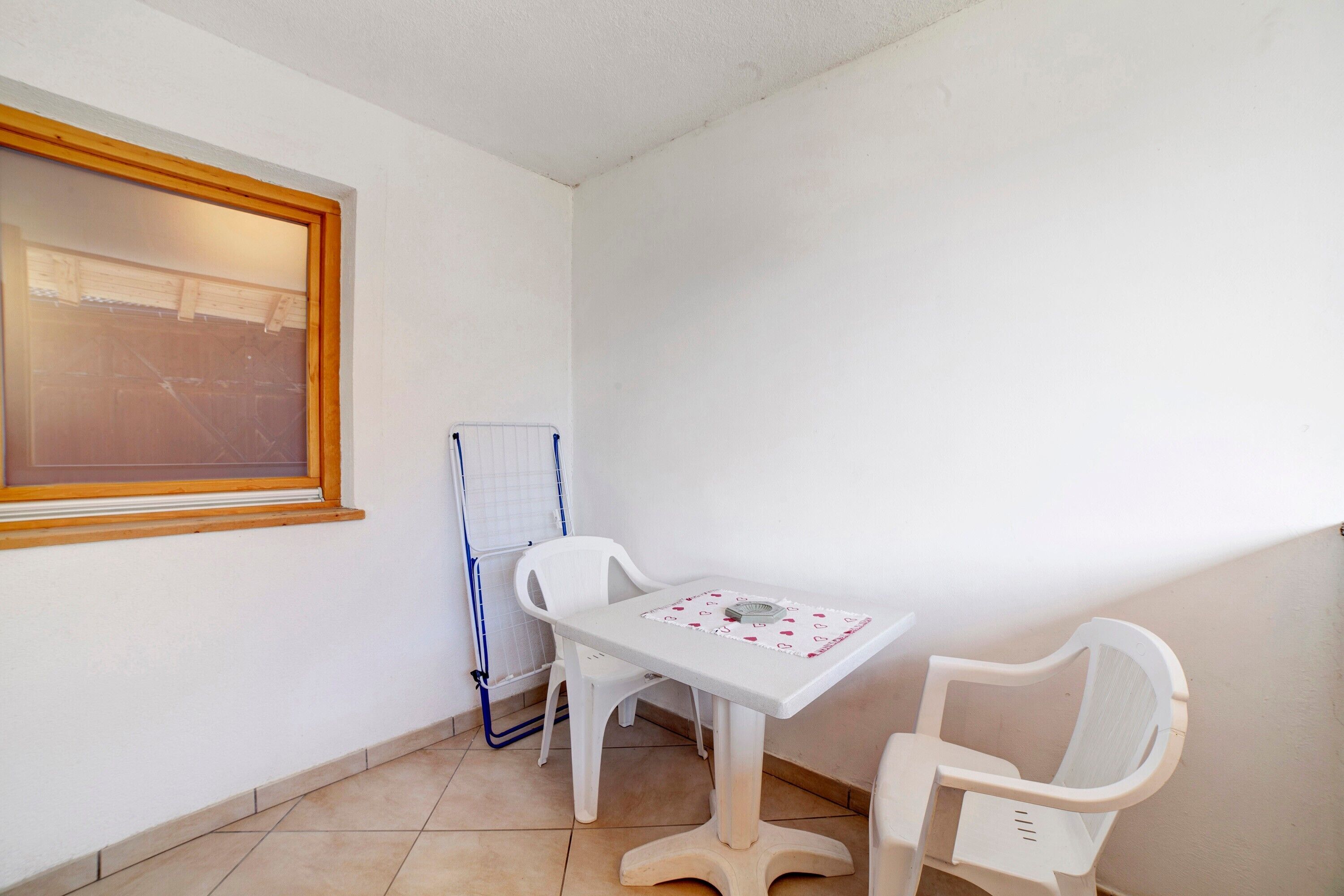 Une petite pièce avec une table blanche et deux chaises blanches, une fenêtre avec un cadre en bois, et un objet bleu appuyé contre le mur.