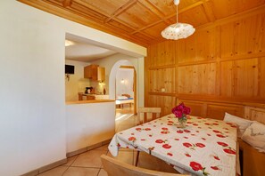 A room with a wooden ceiling, a bed with a floral bedspread, a table with a vase of flowers, and a view into the kitchen.