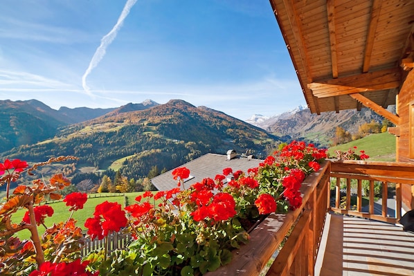 A wooden balcony with red flowers, a mountain view, and a clear sky.