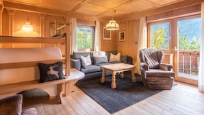 A cozy room with a sofa, a chair, a table, and a bench. There are windows with a view of trees and mountains.