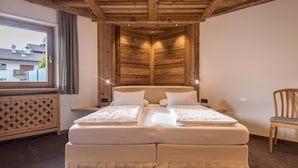 A bedroom with a wooden ceiling, a large bed with white linens, a window with curtains, and a wooden chair.