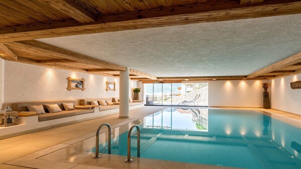 A pool area with a wooden ceiling, a seating area with cushions, and a view of the outside.
