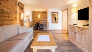 A modern living room with a sofa, a wooden coffee table, a TV, and a kitchen area in the background. - Apartment 'Cesa Sassela 2' with Mountain View, Sauna, Pool, Garden & Wi-Fi (Ortisei)