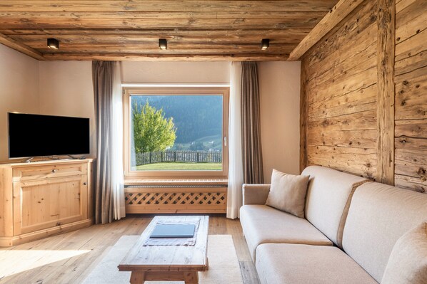 A living room with a wooden ceiling, a television on a wooden cabinet, a beige sofa, and a window with a view of greenery and mountains.