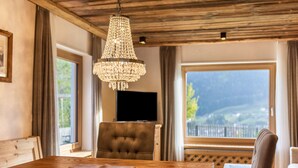A dining area with a wooden table, a chandelier, and a view of a mountainous landscape through large windows. - Apartment 'Cesa Sassela Relax' with Mountain View, Sauna, Pool, Garden, Balcony & Wi-Fi (Ortisei)