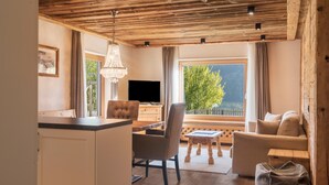 A cozy living room with a wooden ceiling, a television, a dining area with a table and chairs, and a large window with a view of greenery. - Apartment 'Cesa Sassela Relax' with Mountain View, Sauna, Pool, Garden, Balcony & Wi-Fi (Ortisei)