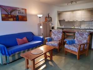 A living room with a sofa, two patterned armchairs, a wooden coffee table, a lamp, and a kitchen area with cabinets and a microwave.
