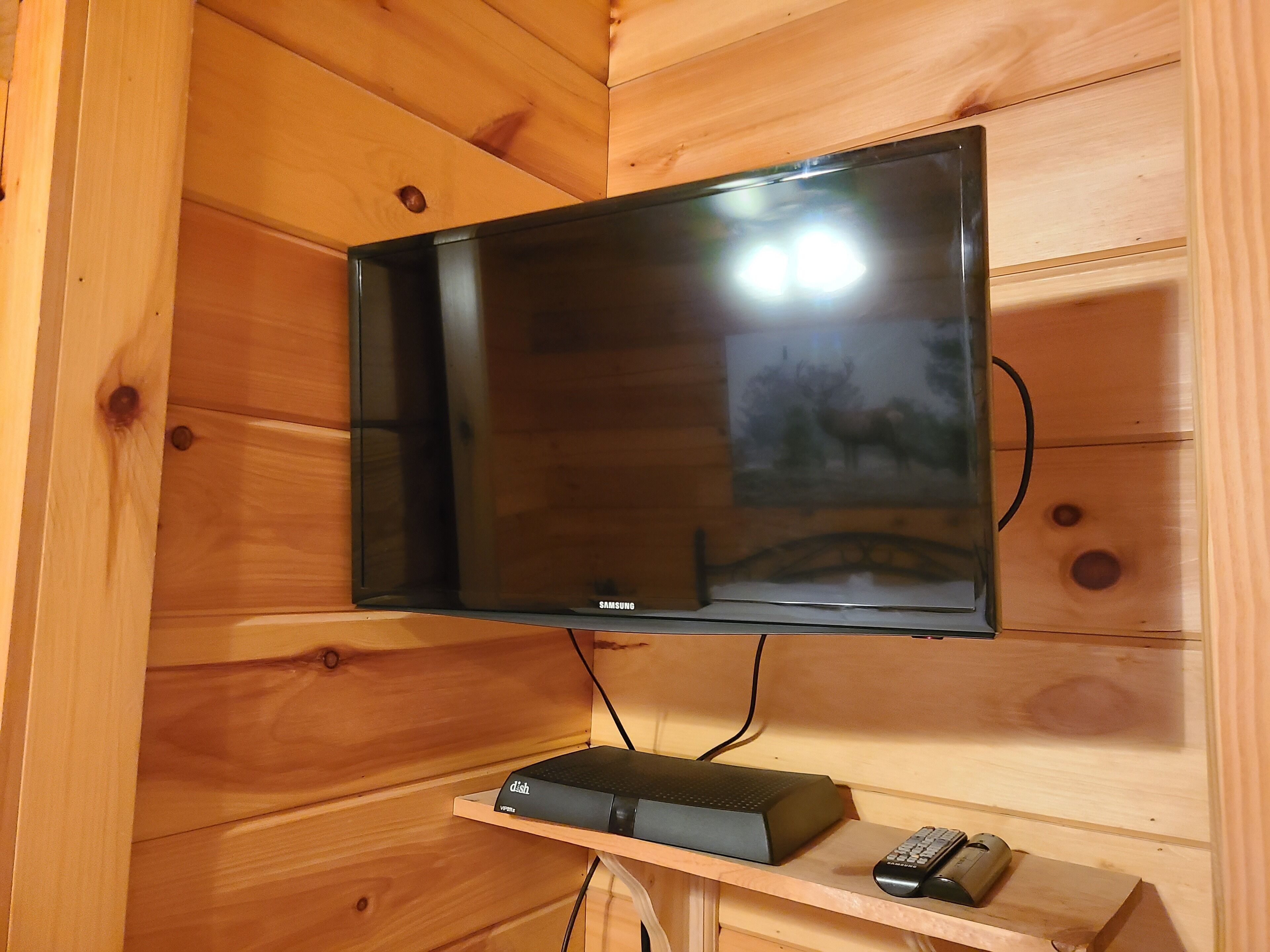 A flat-screen TV mounted on a wooden wall, with a shelf below holding a DVD player and a remote control.