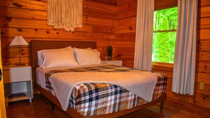 A wooden cabin bedroom with a bed, nightstand, lamp, and window with curtains.