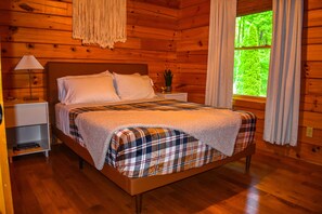 Quarto de cabana de madeira com cama, mesa de cabeceira, abajur e janela com cortinas.