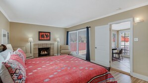 A bedroom with a large bed, a fireplace, a balcony with a table and chairs, and a door leading to an outdoor area.