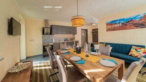 Un salon moderne avec une table à manger pour quatre personnes, un canapé, un téléviseur fixé au mur et une cuisine en arrière-plan.
