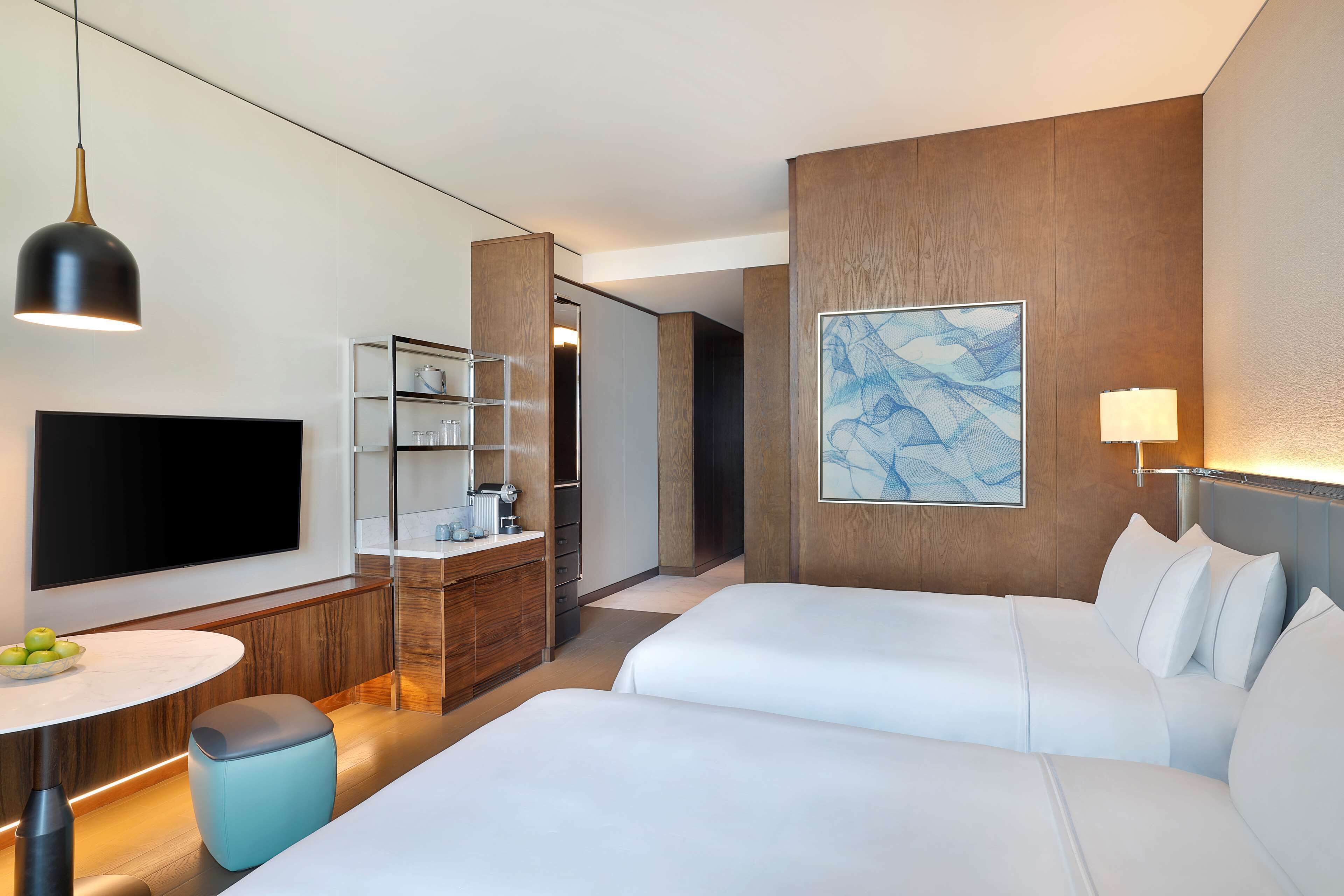 A modern hotel room with a flat-screen TV, a desk with a lamp, a minibar, and a large abstract painting on the wall.