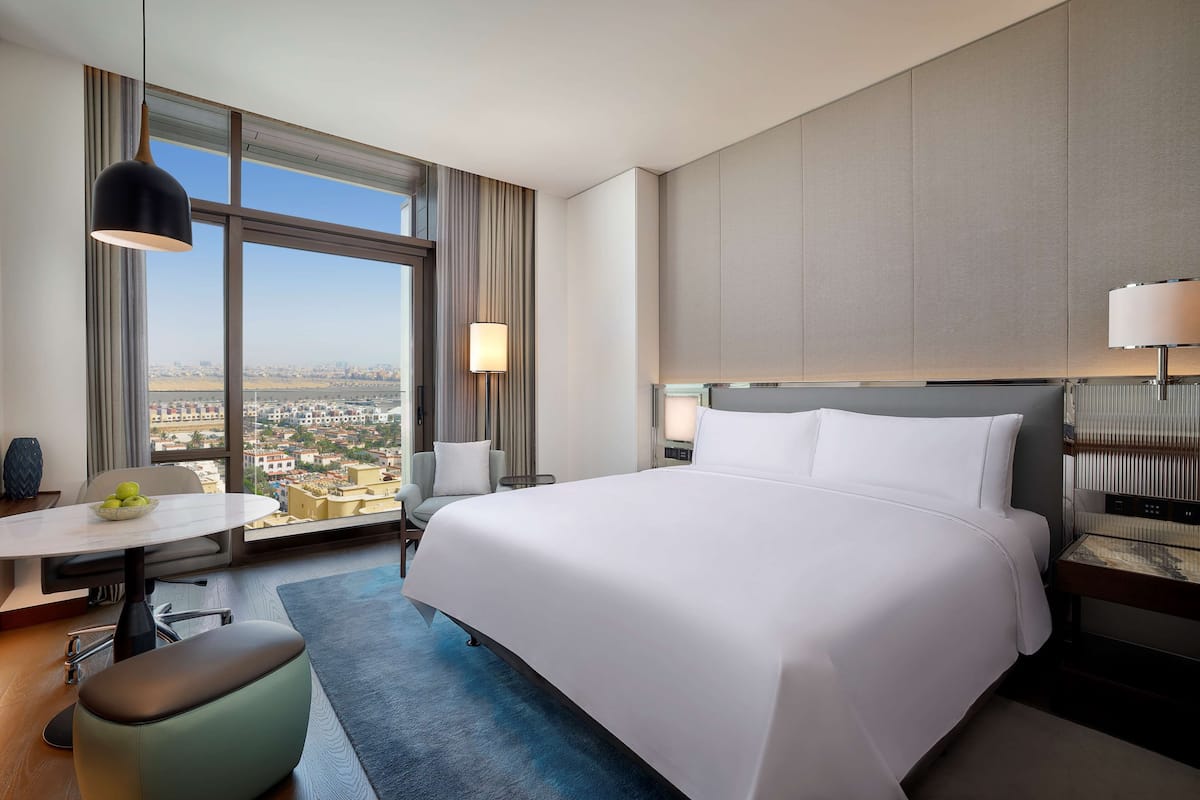 A modern hotel room with a large bed, a desk with a chair, a dining table, and a view of the city through large windows.