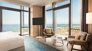 Ein Hotelzimmer mit einem großen Bett, zwei Sesseln, einem Couchtisch und einer Stehlampe. Das Zimmer verfügt über große Fenster mit Blick auf das Meer und den Strand.