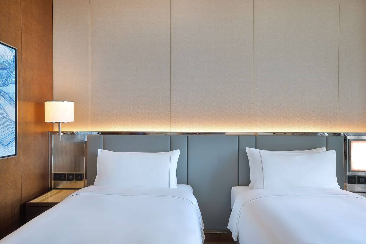 A hotel room with two beds, a nightstand with a lamp, and a large window.
