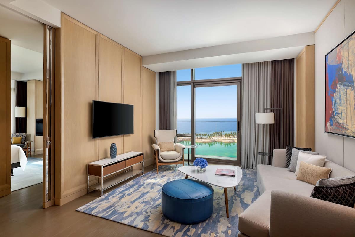 A modern hotel room with a large window, a flat-screen TV, a sofa, a coffee table, and a view of the ocean.