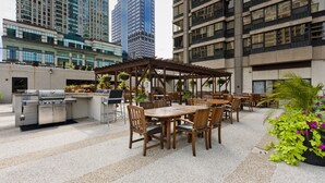 Ein Terrassenbereich im Freien mit Grill, Esstischen und Stühlen, umgeben von Hochhäusern.