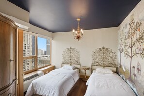 Ein Schlafzimmer mit zwei Betten, einer Chandelieber, einem großen Fenster mit Blick auf Gebäude und einem Wandgemälde.