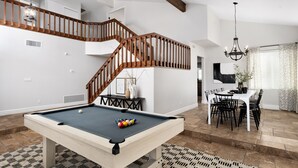 A modern living room with a pool table, a wooden staircase, and a dining area.