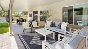 A covered patio area with a white sofa, white chairs, a coffee table, and a dining table.