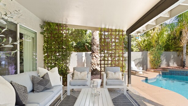 A covered patio area with a pool, a white sofa with patterned cushions, two white armchairs, and a coffee table.