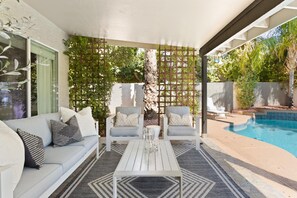 Un patio couvert avec une piscine, un canapé blanc agrémenté de coussins à motifs, deux fauteuils blancs et une table basse.