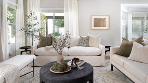A modern living room with a sofa, coffee table, and a bench. There is a window with curtains, a framed picture on the wall, and a small table with a vase and decorative items.