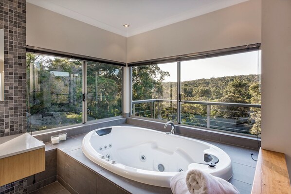 A modern bathroom with a large bathtub, a glass-enclosed shower, and a view of a forested hillside.