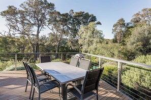 An outdoor dining area with a table and chairs, surrounded by a wooden deck and metal railing, overlooking a natural landscape with trees and shrubs.