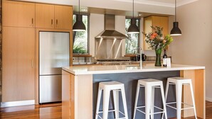 Una cocina moderna con armarios de madera, un refrigerador de acero inoxidable, una isla central con taburetes de bar, iluminación colgante y vistas a la vegetación exterior.