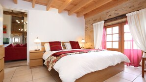 Un dormitorio con una cama grande, vigas de madera, pared de piedra y vistas al exterior a través de una ventana con cortinas.