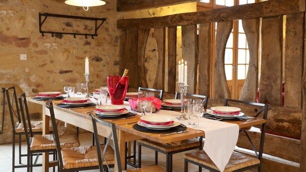 Un comedor rústico con una larga mesa de madera dispuesta para una comida, paredes de piedra y vigas de madera.