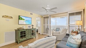 A living room with a TV, a ceiling fan, a green cabinet, a sofa, and a window with blinds. - 180° Gulf Views | 1st Floor Condo| Pool & Walk to Dining (Fort Myers Beach)
